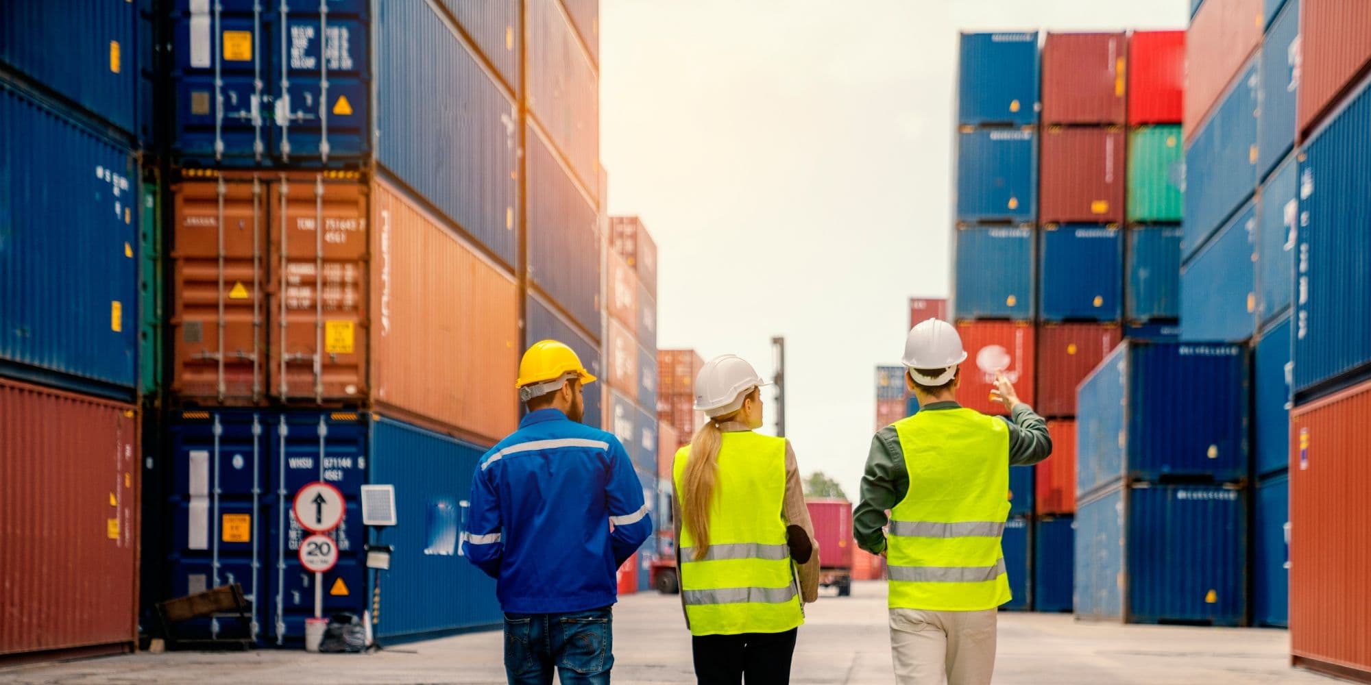 Drei Menschen laufen durch einen Containerhafen. Sie tragen Helme und Sicherheitswesten