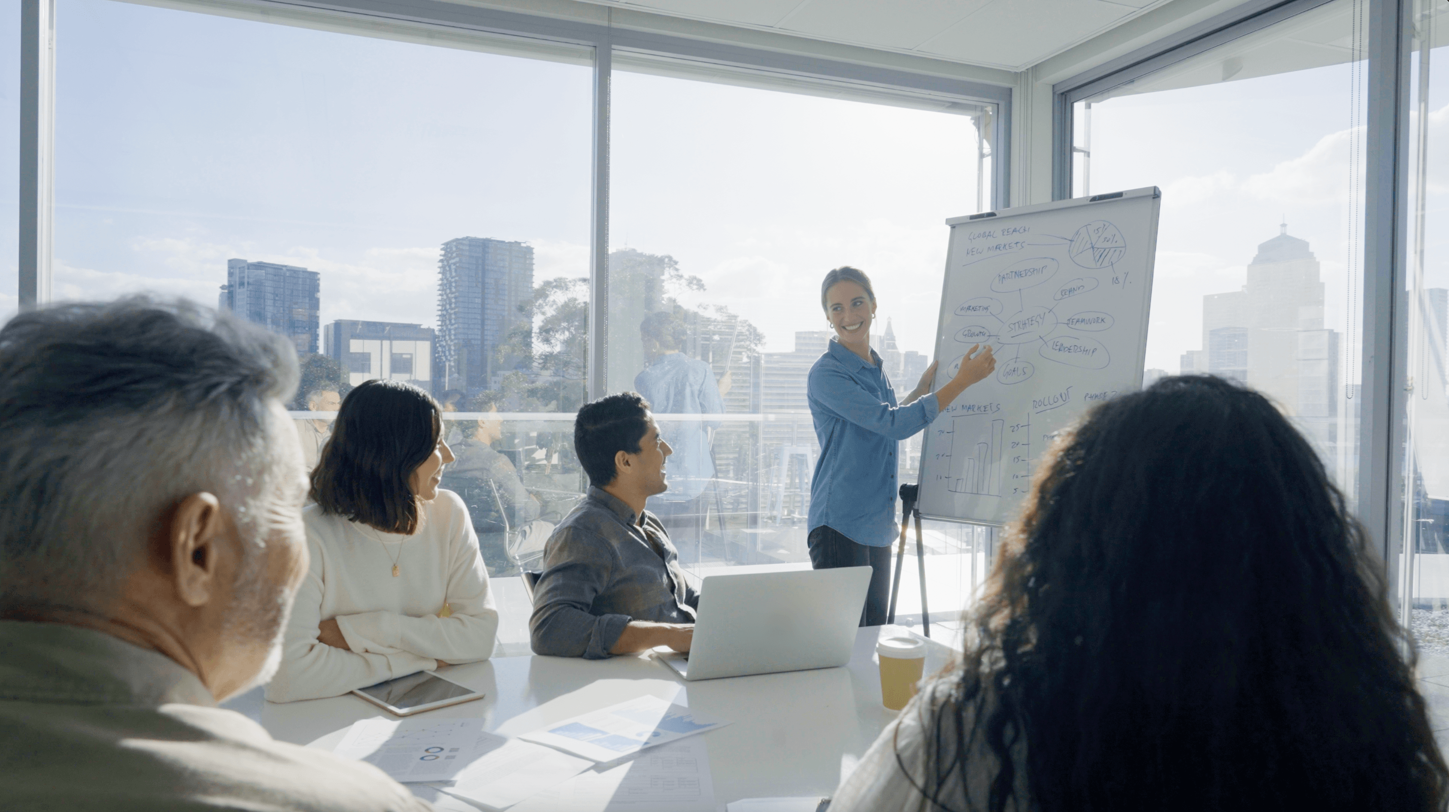 Die Sonne scheint in den Meetingraum. Eine Frau präsentiert mithilfe eines Whiteboards.