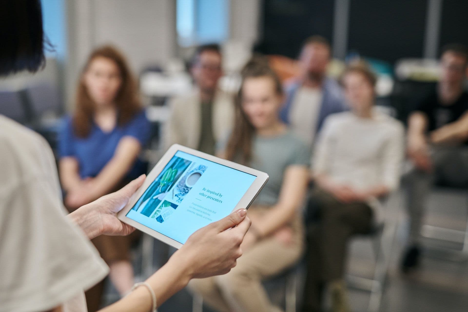 Im Fokus steht ein Tablet mit dem Slogan 'Be inspired by other presenters'. Im Hintergrund sitzen Menschen und hören zu.